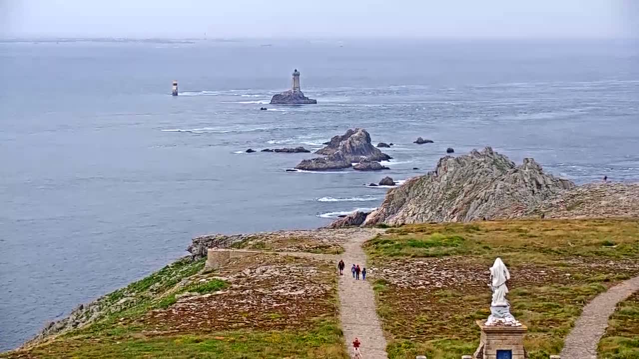 A coastal view shows a rocky shoreline, a lighthouse on an island in the distance, several people walking on a path, and a statue on a pedestal.