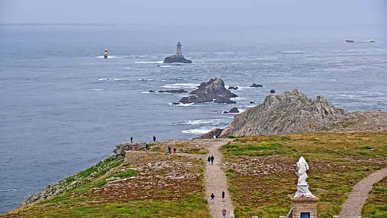 A coastal overlook on an overcast day shows a rocky shoreline, a lighthouse in the distance, and several people walking along a path toward a statue.