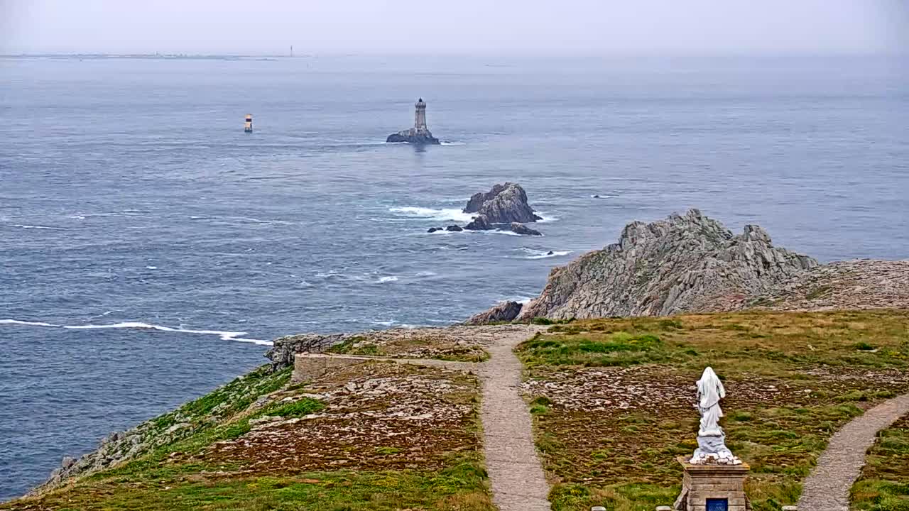 A coastal overlook with a statue shows a hazy ocean view of rocky islands and distant lighthouses under an overcast sky.