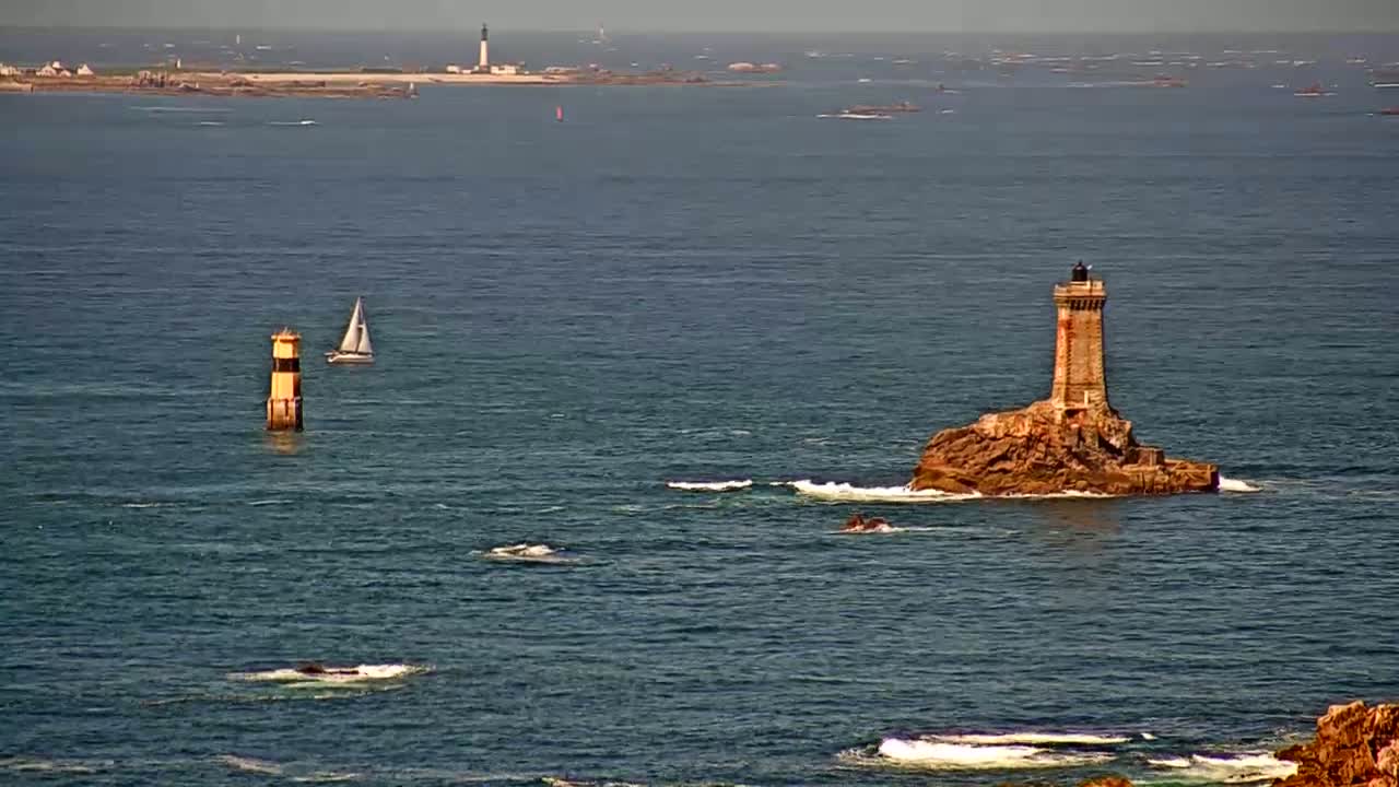 A sailboat sails past a small, yellow lighthouse in the foreground, while a larger lighthouse sits on a rocky outcrop in the middle ground, with a distant island and lighthouse visible on the horizon under calm, sunny conditions.