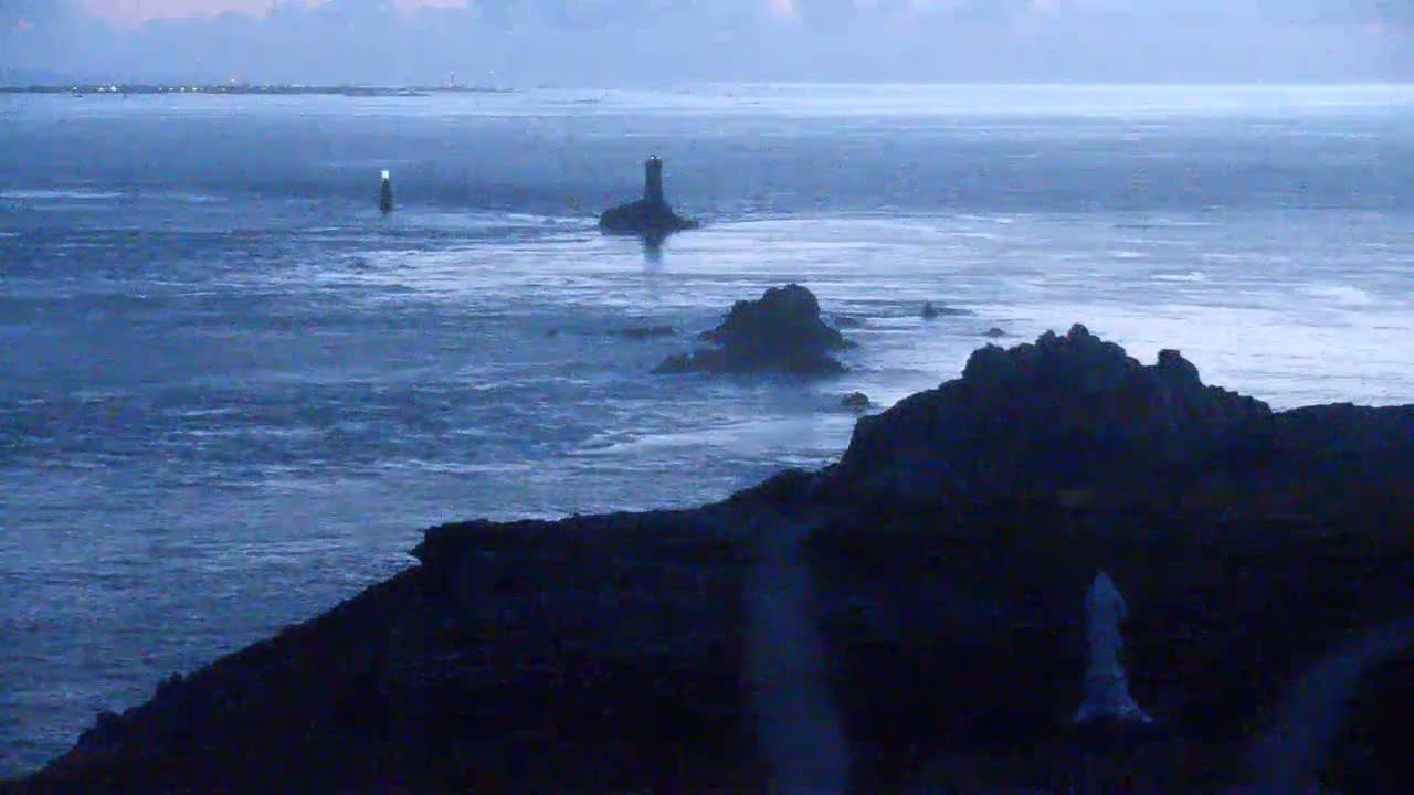 Two lighthouses stand in a calm, gray sea under a hazy sky, with a distant landmass visible in the background.