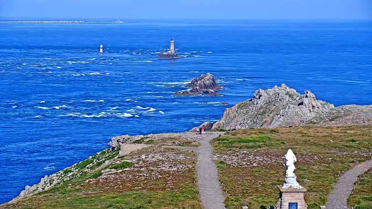 A coastal view shows a rocky headland with a statue and paths overlooking a blue ocean with several small islands and lighthouses under a clear sky.