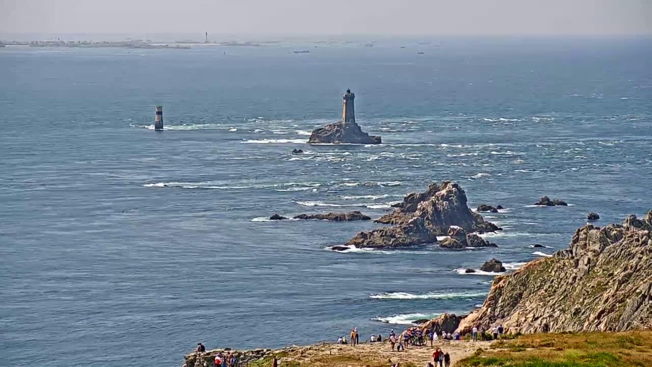 A coastal scene on a hazy day shows a lighthouse on a small island, other rocky outcrops, and a group of people gathered on a cliff overlooking the ocean.