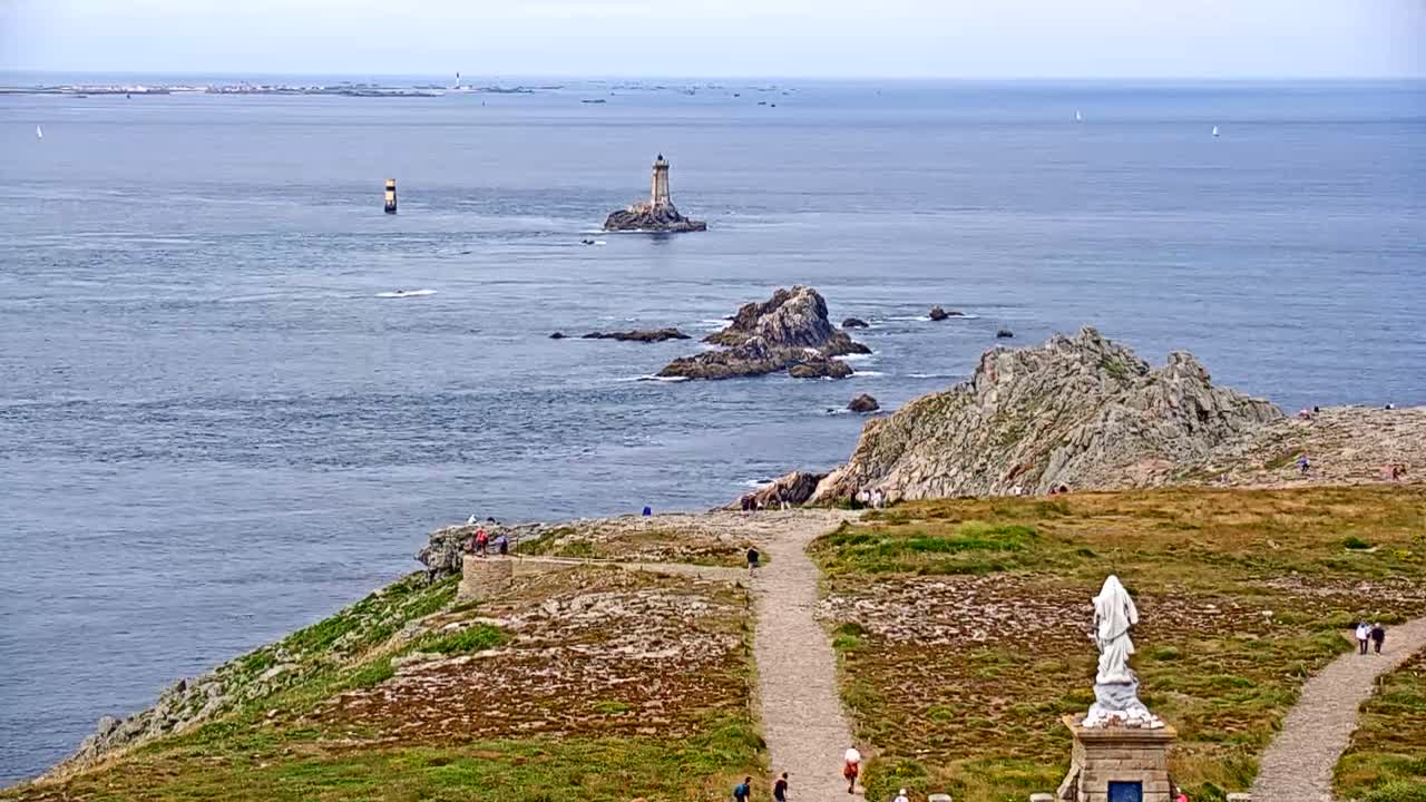 A coastal view on a mostly sunny day shows a lighthouse on a small island, rocky outcroppings in the sea, a statue on a grassy clifftop path with a few people walking by.