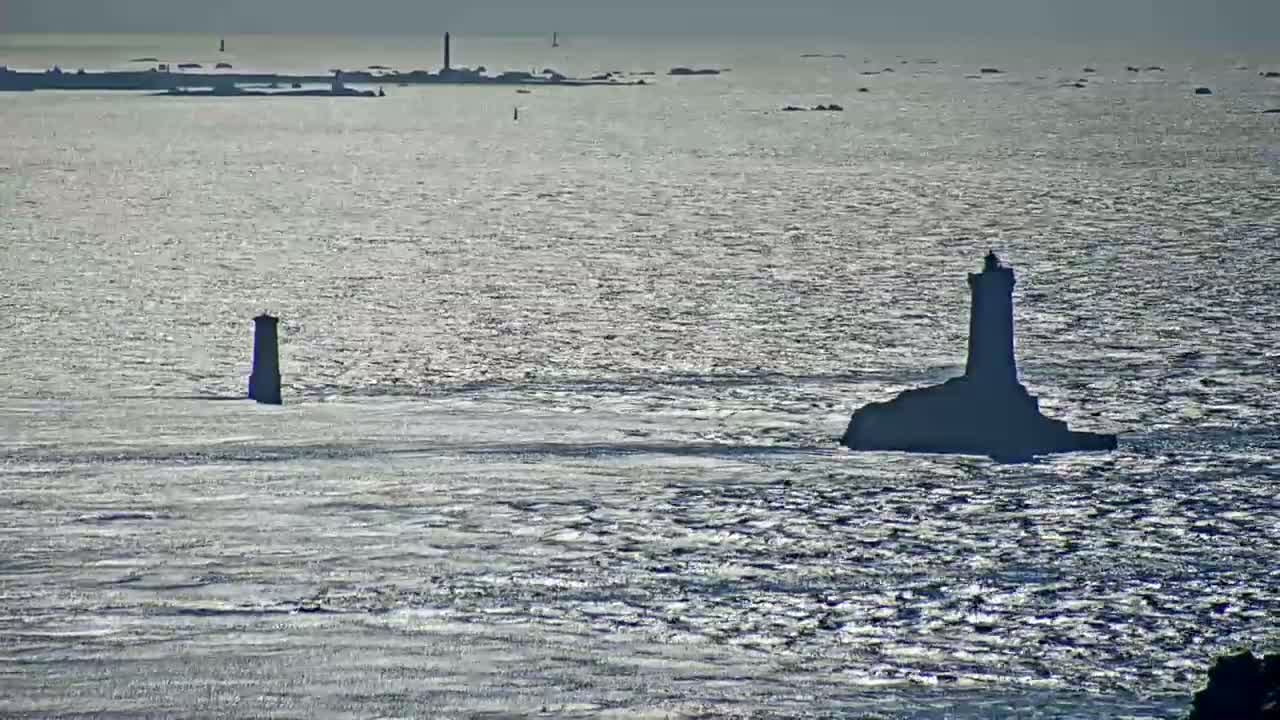 Two lighthouses stand on rocky outcrops in a calm, sunlit sea, with a distant landmass visible in the background.