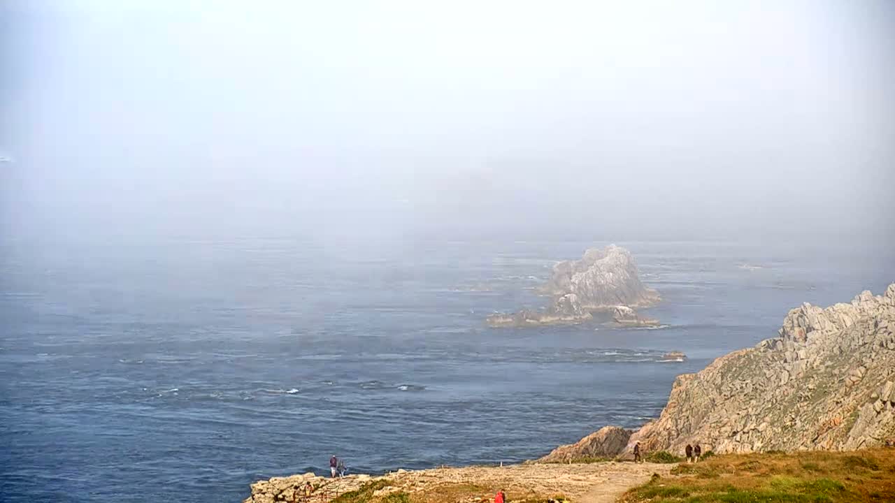 A foggy coastal scene shows a rocky cliff overlooking a blue ocean with several small rocky islands in the distance.