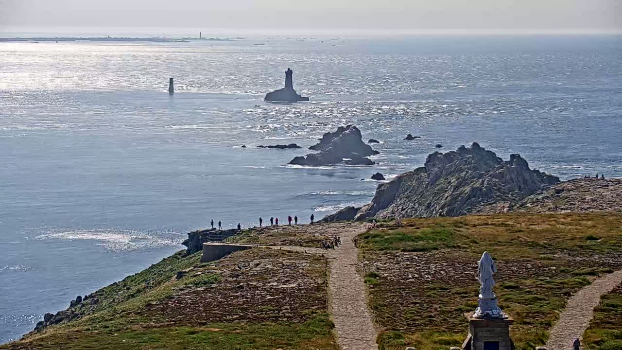 A rocky coastal scene with a lighthouse and a statue visible on a sunny day, overlooking a choppy ocean.
