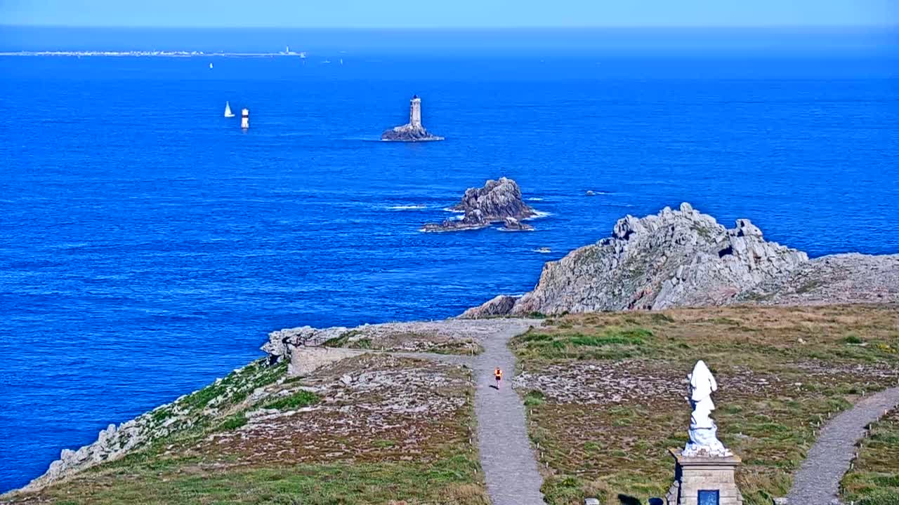 A person walks on a path overlooking a rocky coastline and ocean on a sunny day, with a distant lighthouse and several sailboats visible.