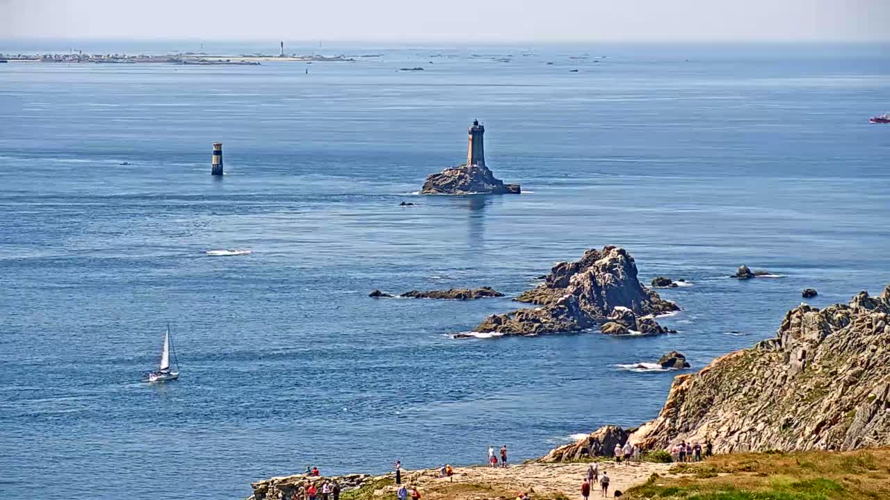 A sailboat and several people are visible on a sunny day, overlooking a rocky coastline and a distant lighthouse on a small island surrounded by blue water.