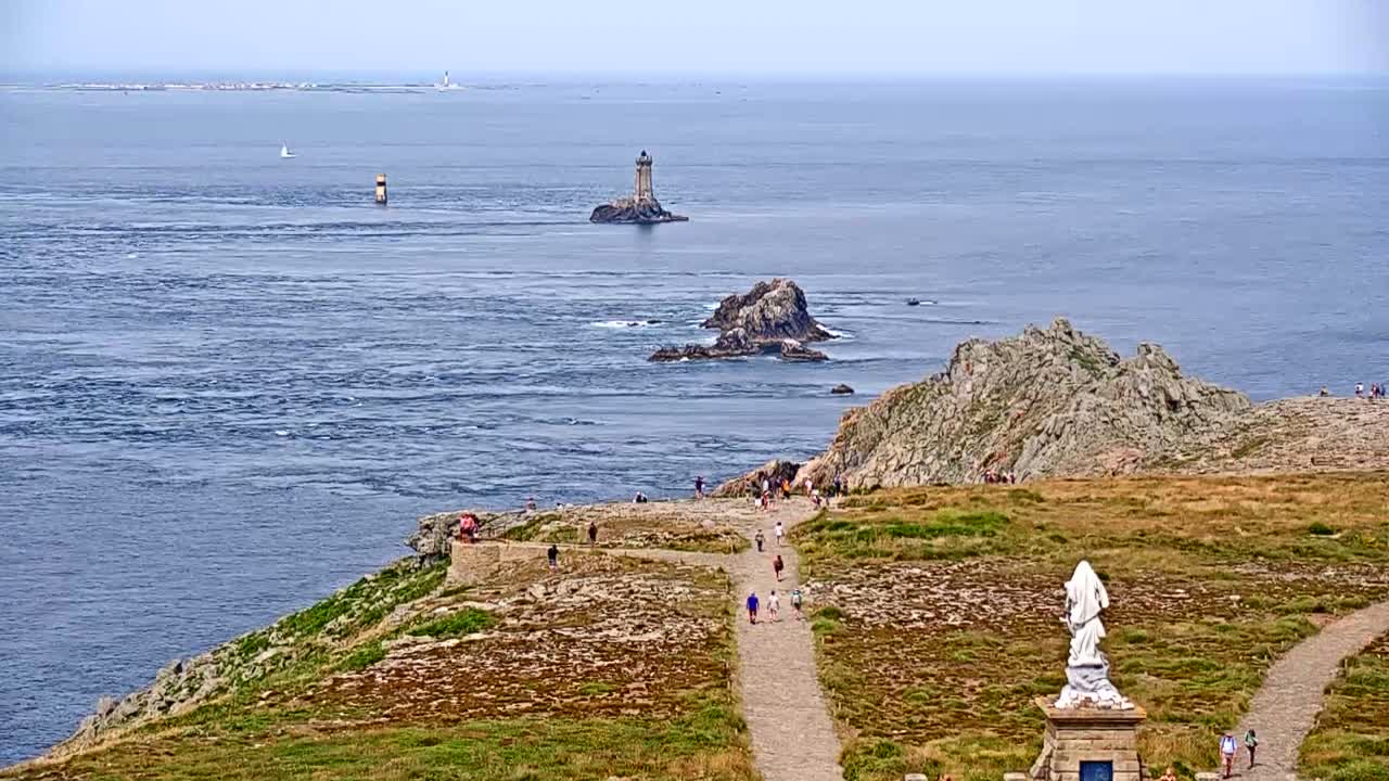 A rocky coastal path with several people walking on it leads to a statue, overlooking a calm ocean with several small islands and a lighthouse visible in the distance.