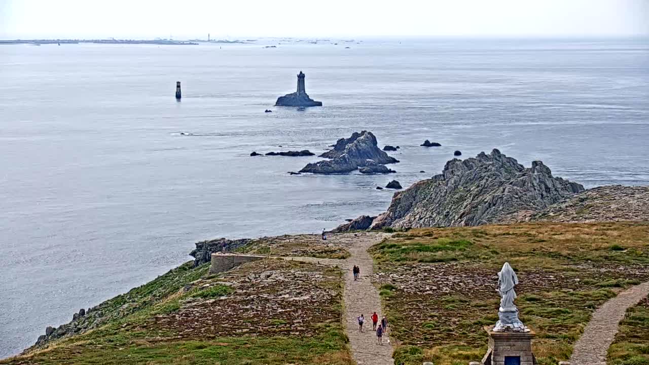 A coastal scene on a hazy day shows a rocky outcrop with a path and a statue, overlooking a body of water with distant lighthouses and islands.