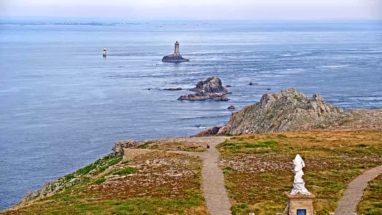 A coastal scene on a calm, hazy day shows a rocky outcrop with a lighthouse in the distance, other smaller rocky islands, and a statue on a grassy cliff overlooking a path.