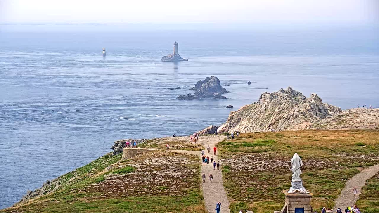 A coastal scene on a hazy day shows a rocky, grassy headland with a statue and people walking on a path overlooking a body of water with several small islands and lighthouses in the distance.