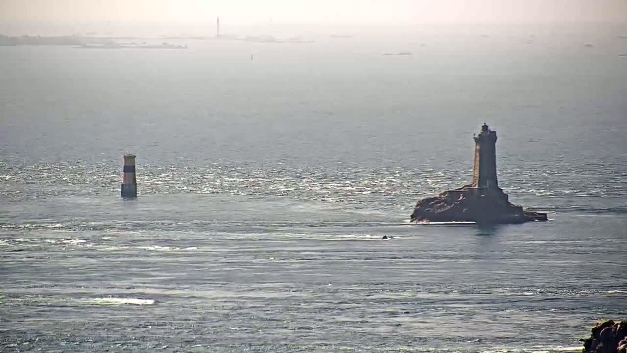 A tall lighthouse sits on a small, rocky island in a calm, hazy sea, with a smaller lighthouse visible in the distance.