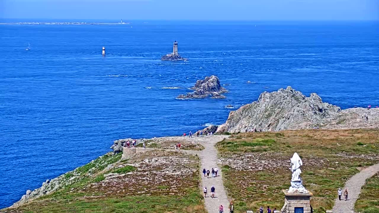 A coastal vista on a clear, sunny day shows a rocky headland with a statue and walking path, overlooking a blue ocean dotted with several lighthouses and small boats.