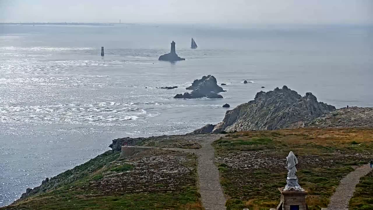 A rocky coastal scene with a statue overlooks a choppy sea dotted with small islands and a sailboat under a hazy sky.