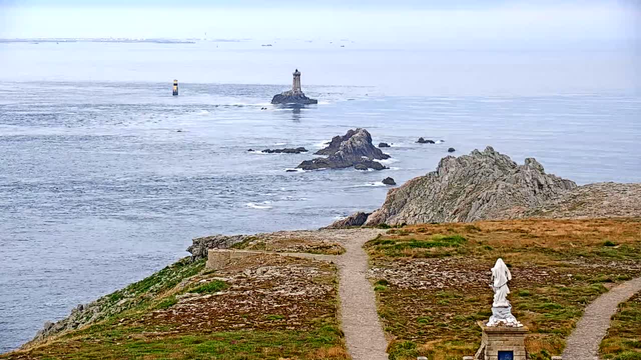 A coastal scene shows a lighthouse on a small island in a hazy ocean, with rocky outcrops nearby, a statue on a grassy cliff, and a path leading to it.