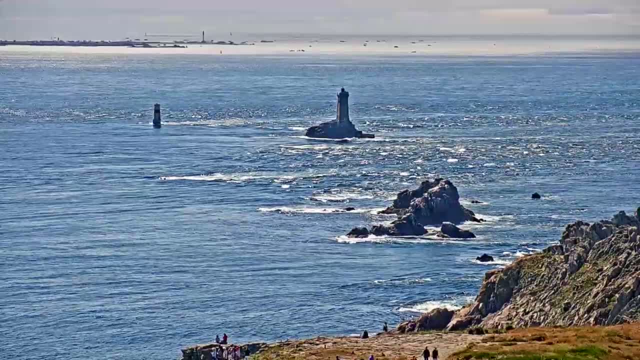A rocky coastline with a lighthouse on a small island in a choppy ocean under a sunny sky.