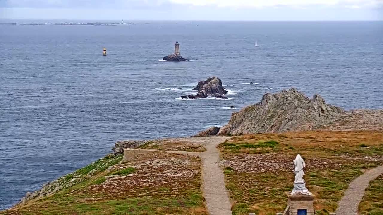A rocky coastal scene under a cloudy sky shows a distant lighthouse, a statue, and a path leading to the ocean.