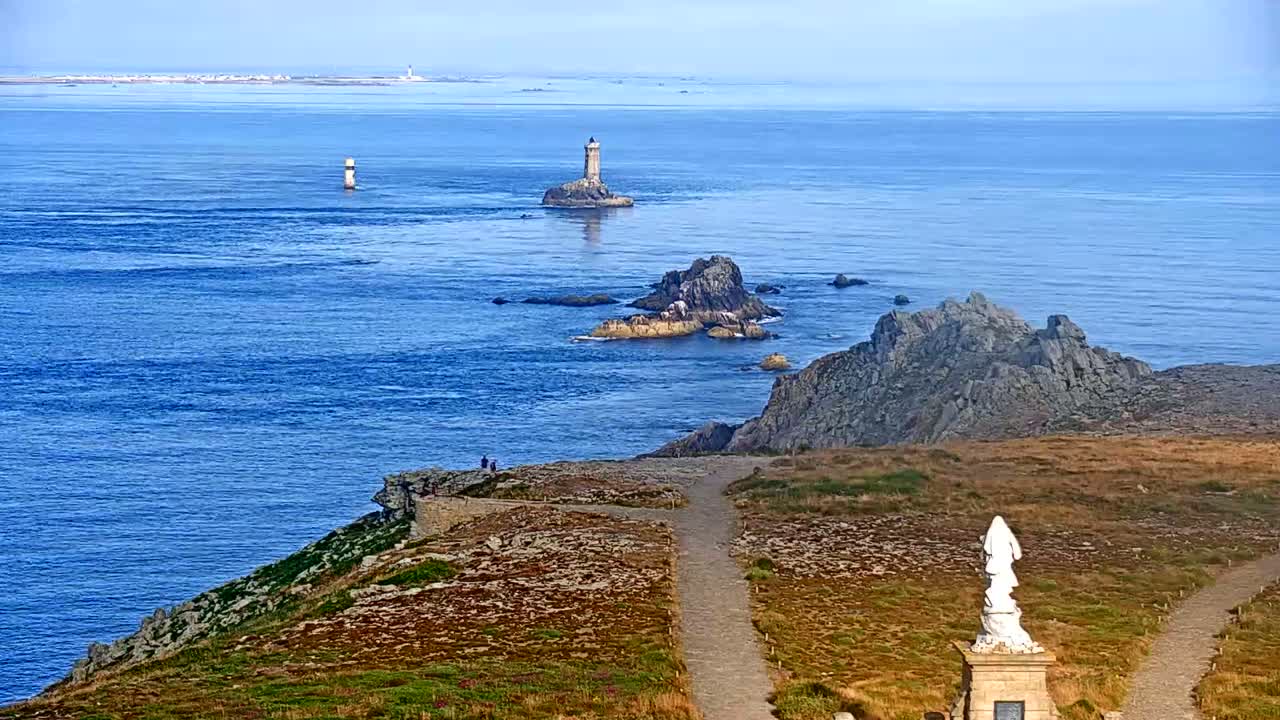 A coastal scene on a clear day shows a rocky shoreline with a statue, paths, and distant lighthouses and islands on a calm ocean.