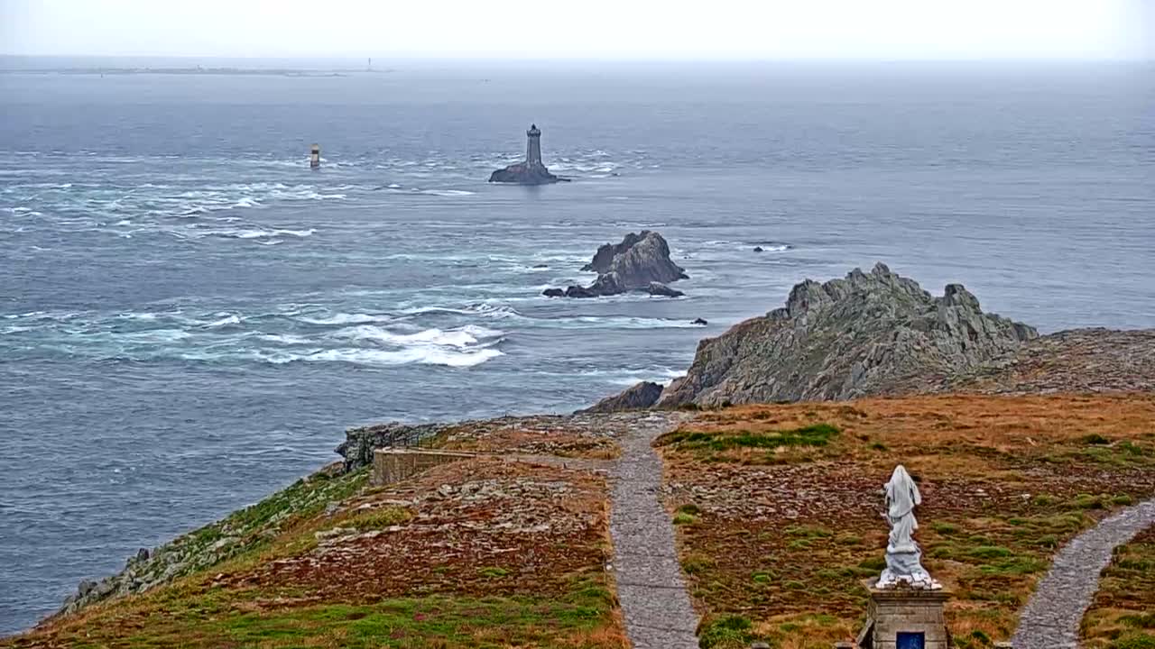 A rocky coastline with a statue and pathways overlooks a choppy sea with several small islands and lighthouses visible in the distance under an overcast sky.