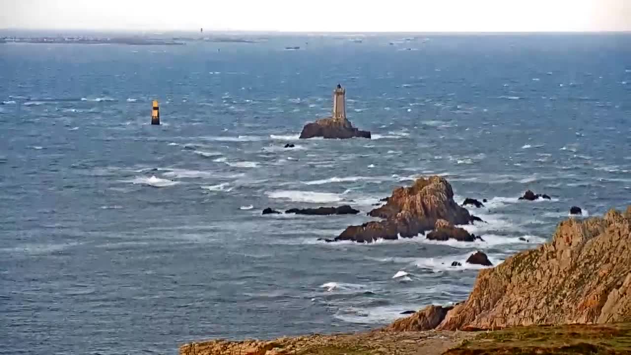 A lighthouse rises from a rocky islet in a windswept, choppy grey-blue sea with a yellow buoy to its left, viewed from a rugged, rocky foreground under a bright overcast sky.