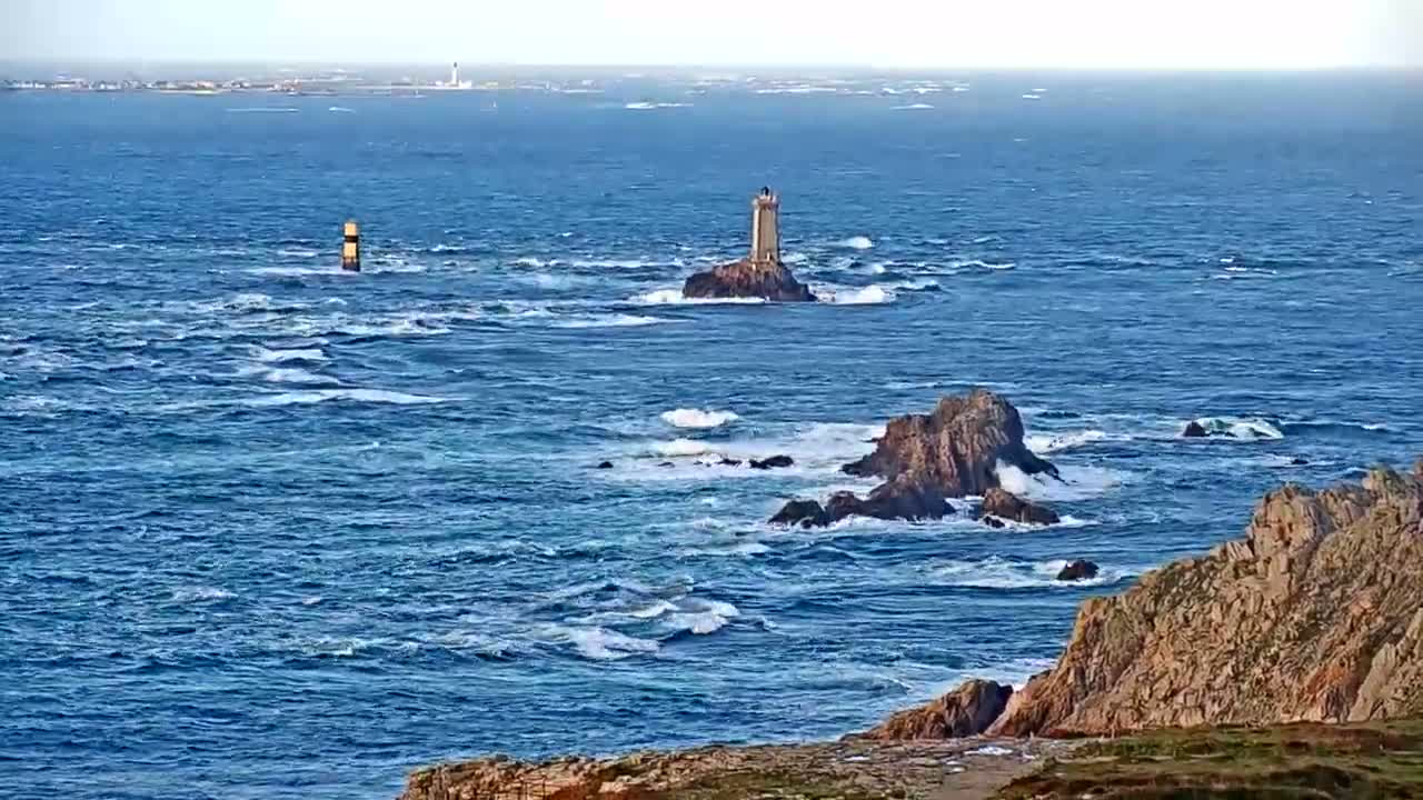 A distant lighthouse stands on a rocky islet amidst choppy blue seas with whitecaps under a bright, hazy sky, flanked by a rugged brown coastline in the foreground.