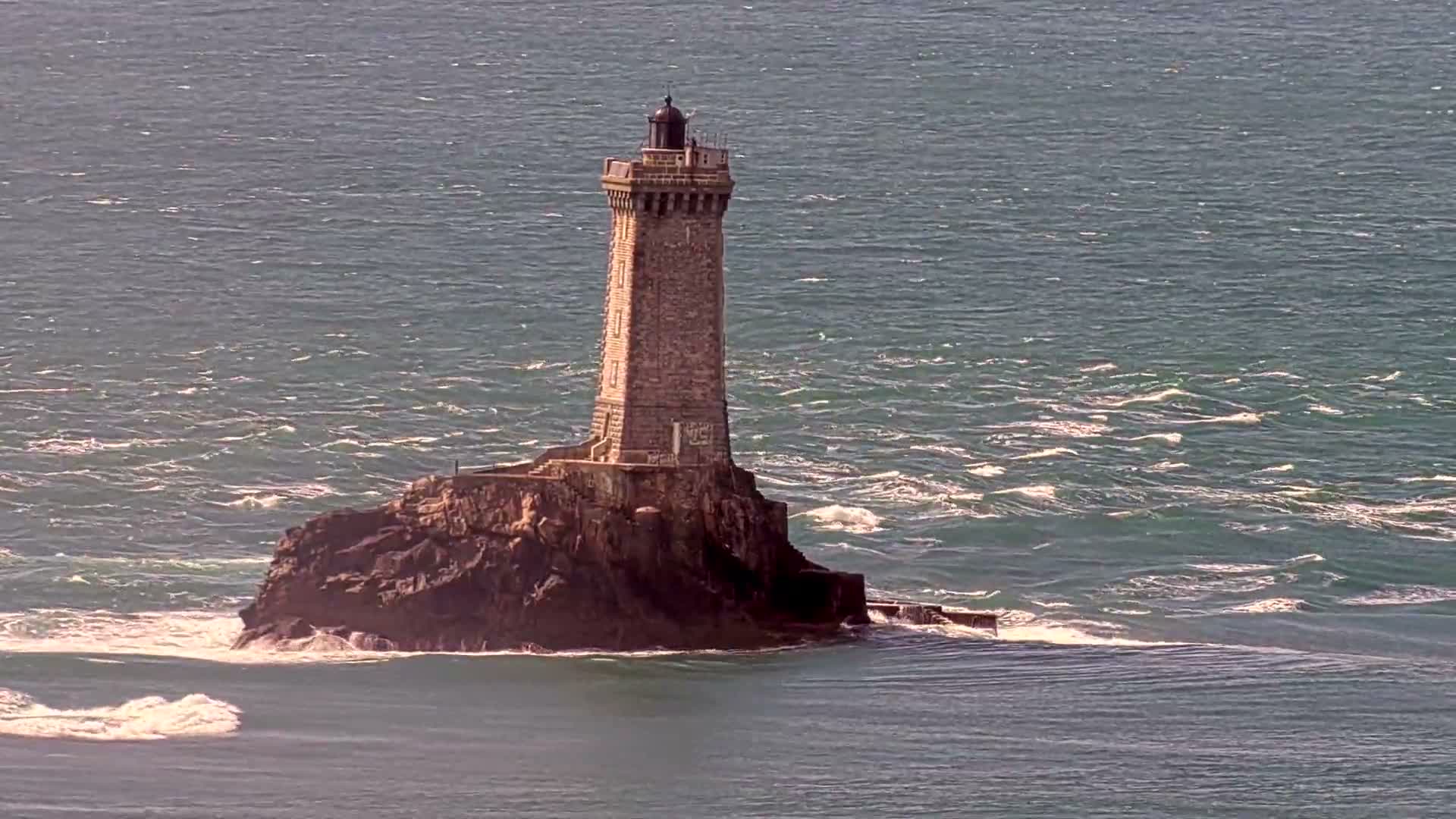 A distant lighthouse stands on a rocky islet amidst choppy blue seas with whitecaps under a bright, hazy sky, flanked by a rugged brown coastline in the foreground.
