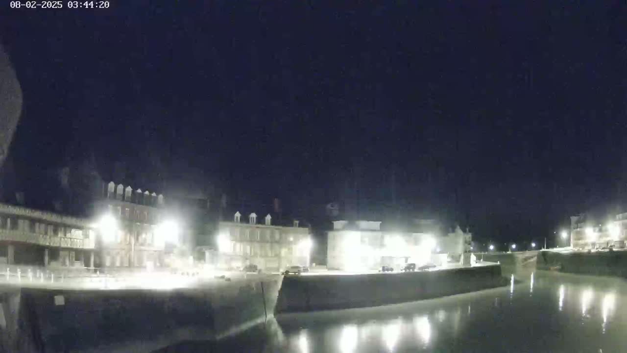 A nighttime view of a calm harbor with illuminated buildings lining the waterfront under a dark sky.