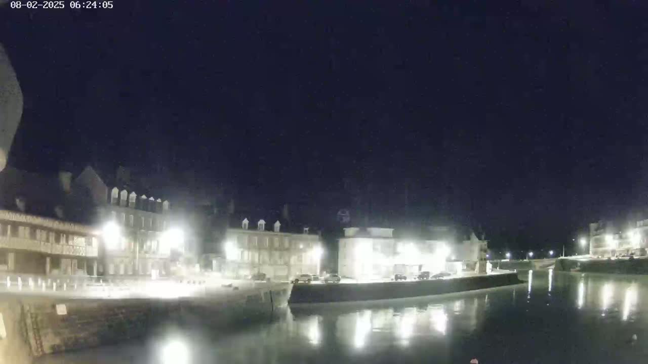 A nighttime view of a calm waterway reflecting illuminated buildings lining its banks.