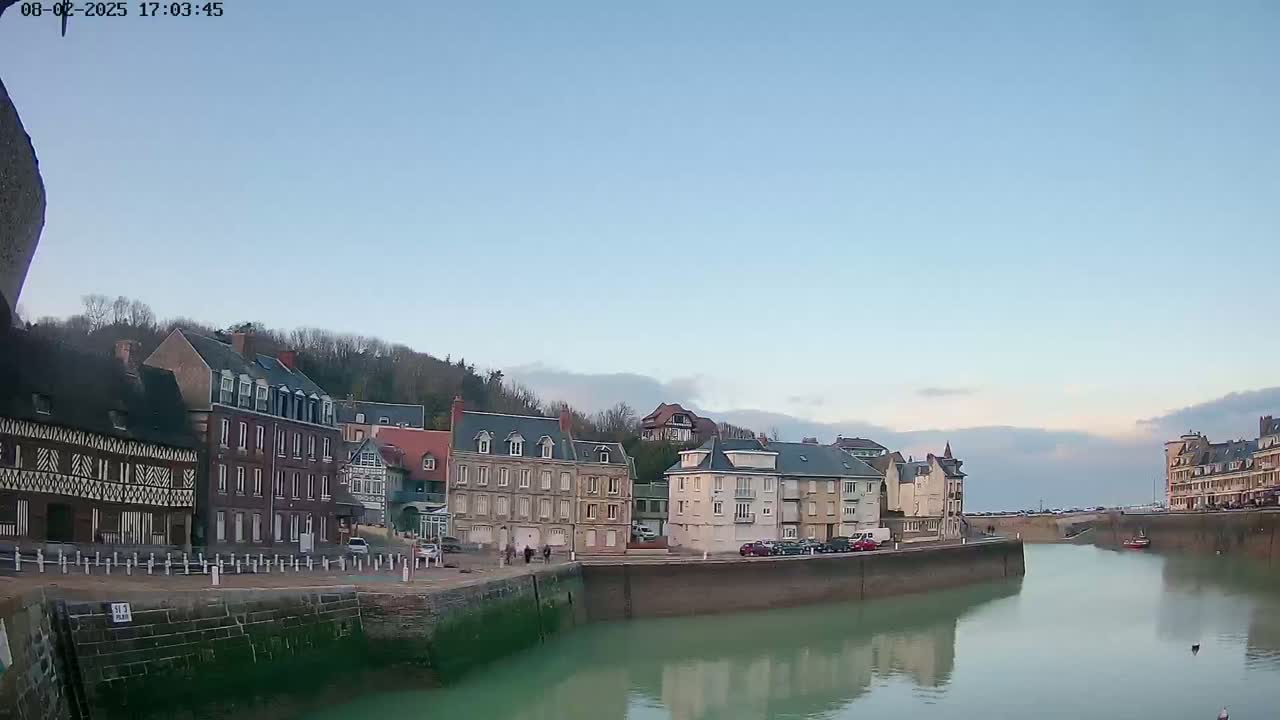 Under a clear, light-blue sky, a calm waterway flows between rows of buildings nestled against a hillside.