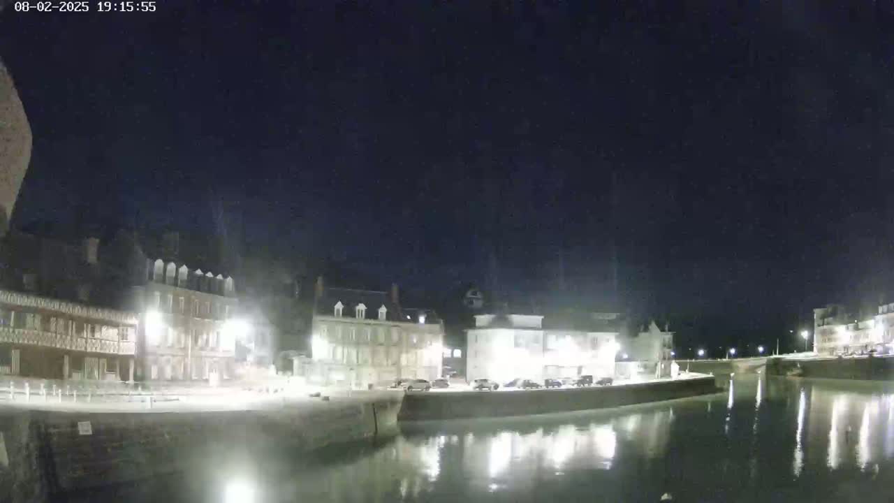 A nighttime view of a calm waterway reflecting illuminated buildings along its banks under a dark sky.