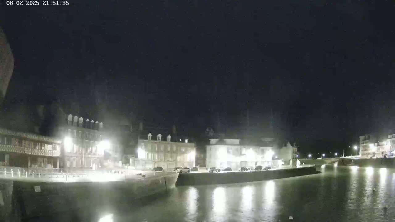 A nighttime view of a calm harbor with several buildings illuminated along the waterfront under a dark sky.