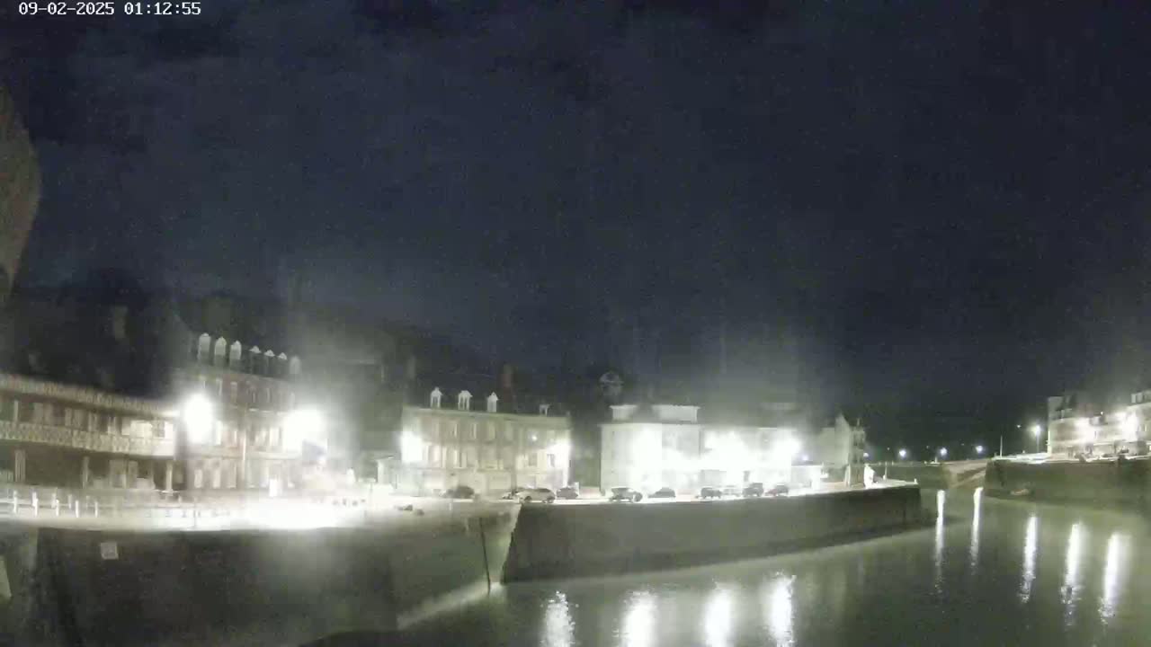 A nighttime view of a calm canal reflecting illuminated buildings along its banks under a dark, clear sky.