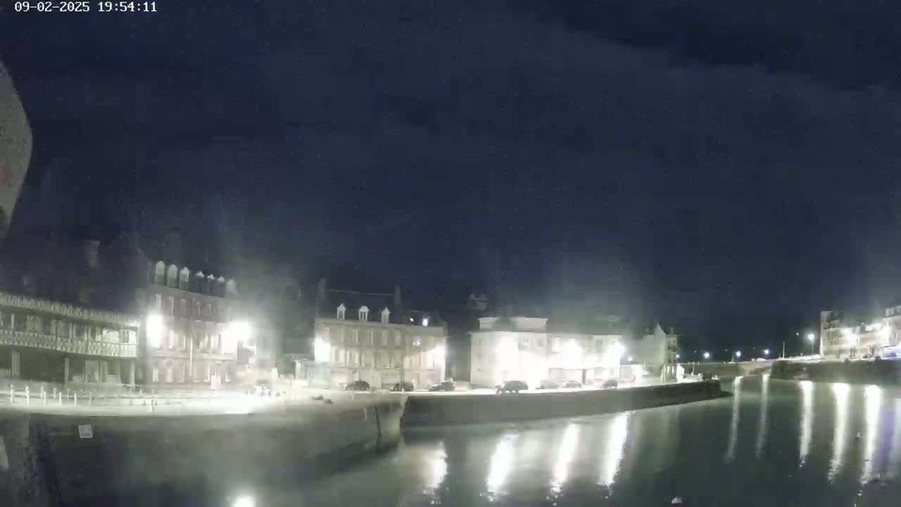A nighttime view of a calm harbor reflecting lights from buildings along its edge under a dark, clear sky.