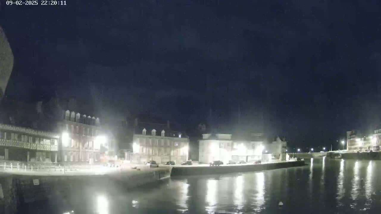 A nighttime view of a waterfront town with calm water reflecting the lights from buildings along the shore under a dark, clear sky.
