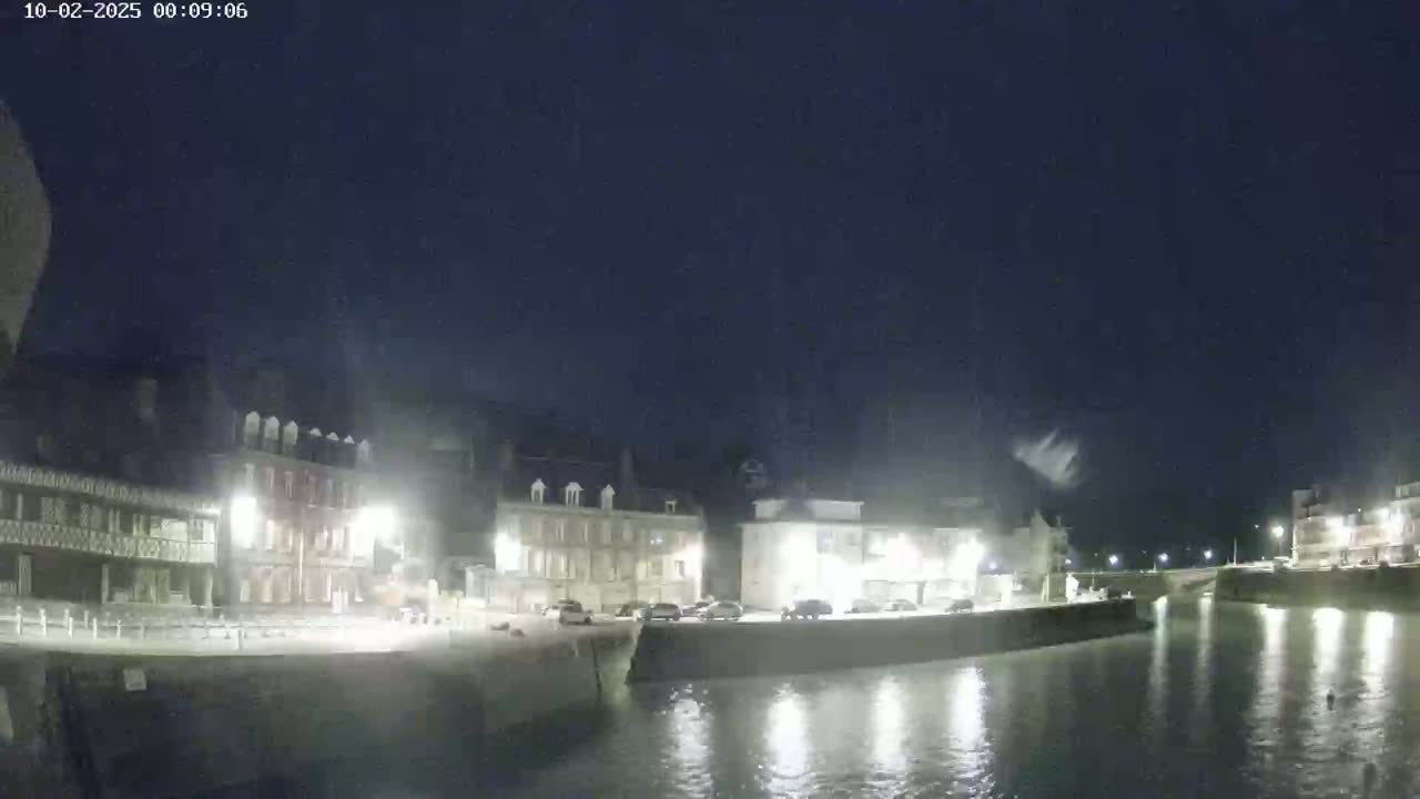A nighttime view of a waterfront town with several buildings lit up along a calm body of water under a dark, clear sky.