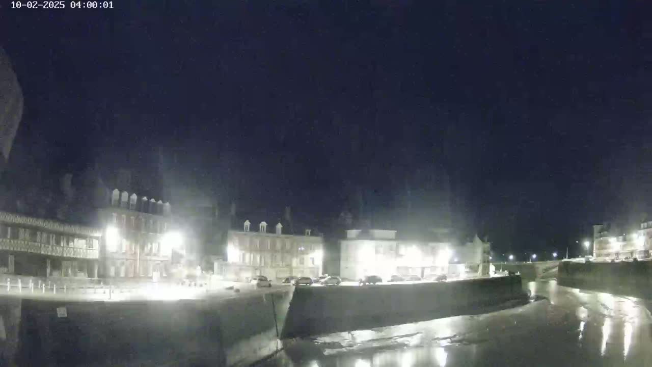 A nighttime view of a town square with several buildings illuminated, overlooking a calm body of water under a clear, dark sky.
