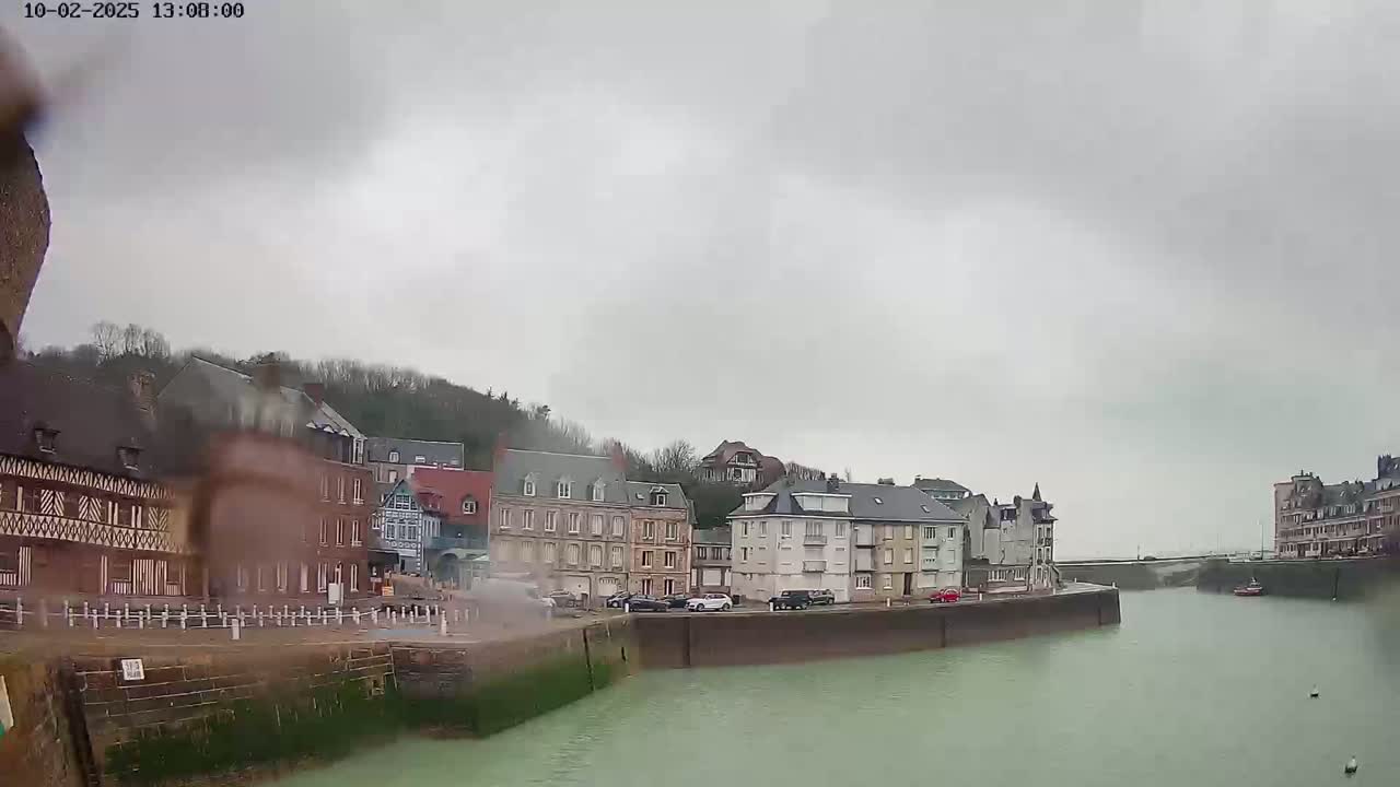 A waterfront town with multi-story buildings lines a calm, green waterway under an overcast sky.