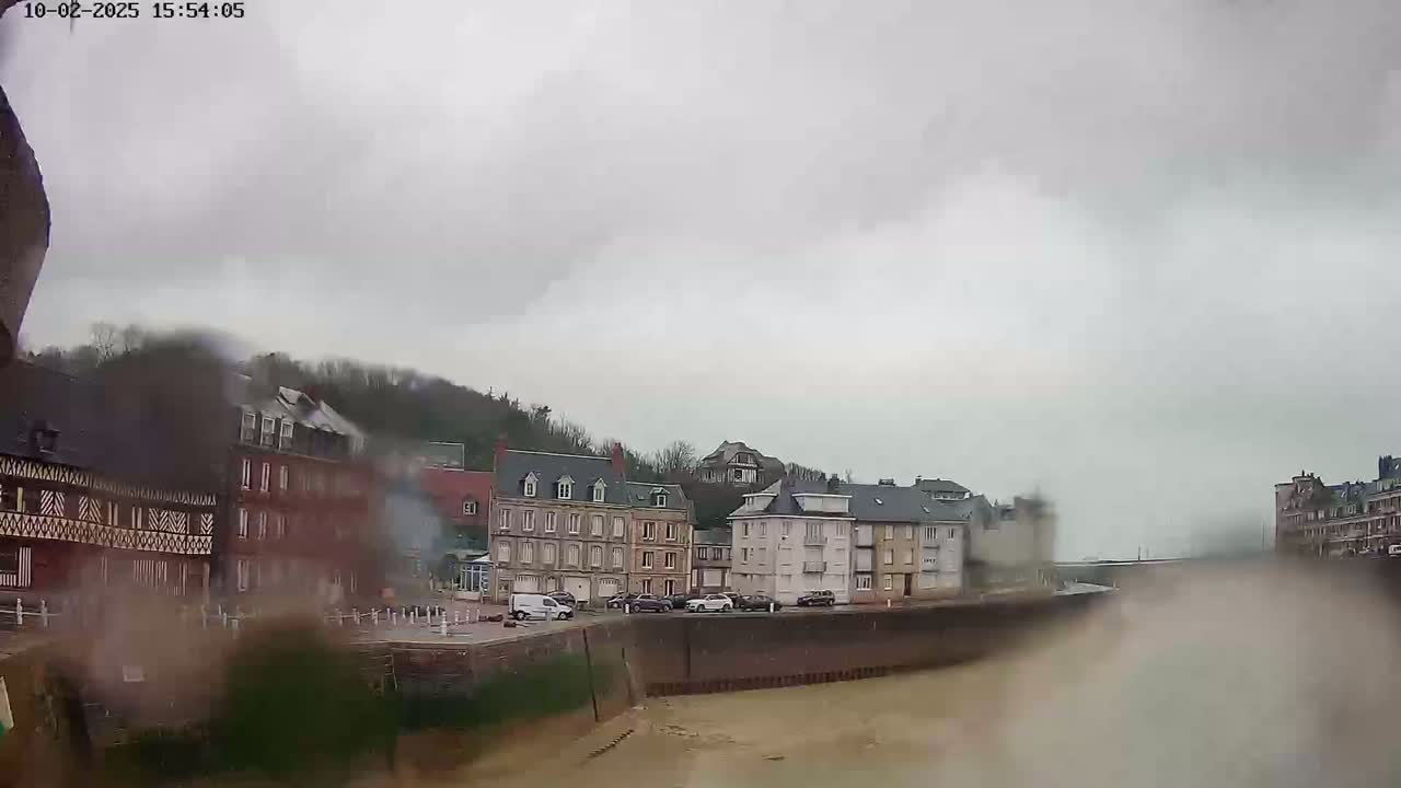 A rain-covered view of a waterfront town with several multi-story buildings, a parking lot, and a calm body of water.