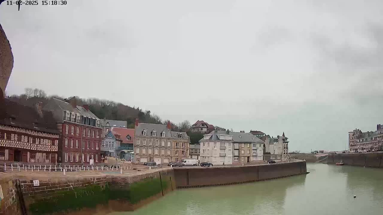 A calm, overcast day reveals a waterfront scene of several multi-story buildings lining a waterway, with a few cars parked nearby.