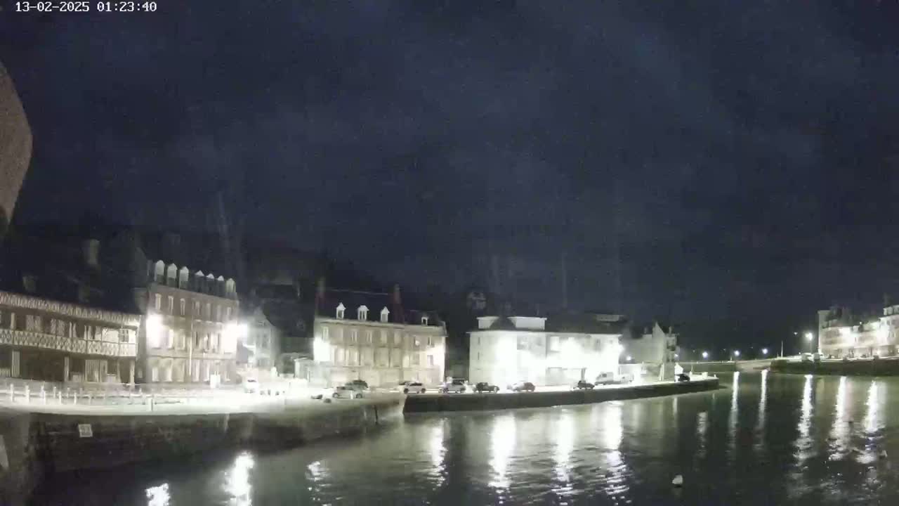 A nighttime view of a waterfront town with buildings lining a calm canal under a dark, cloudy sky.