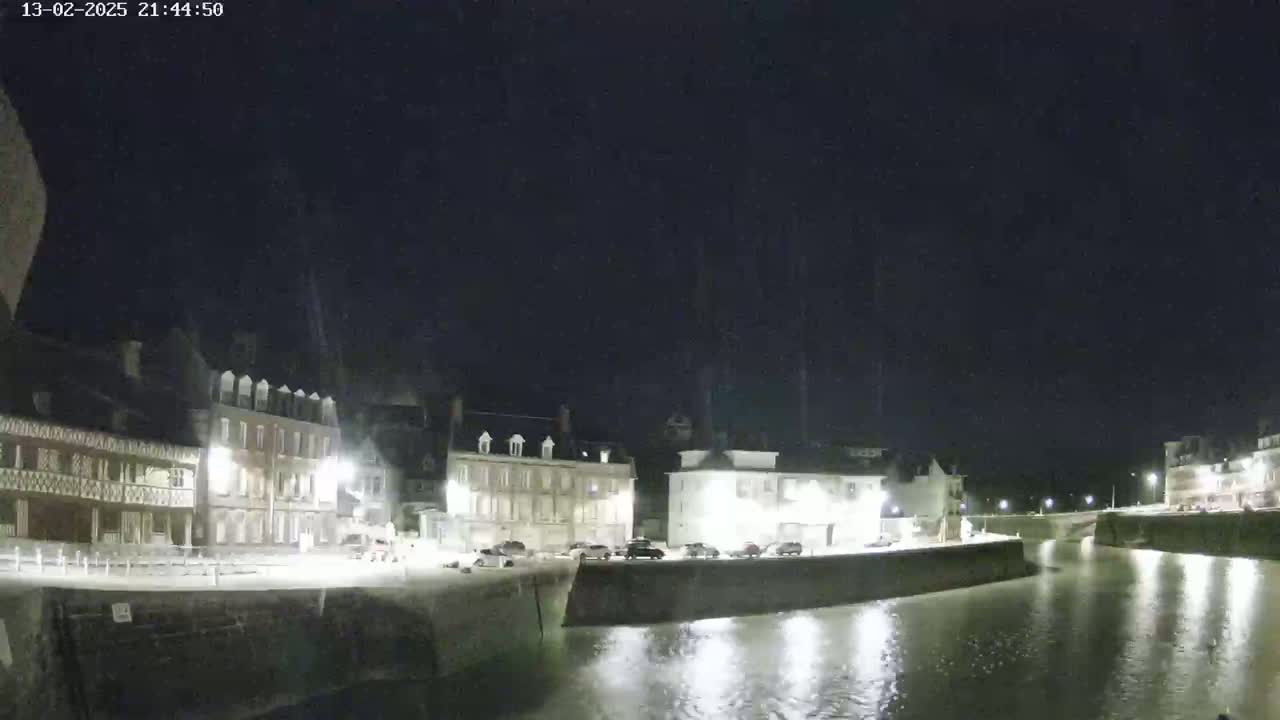 A nighttime view of a waterfront town with several buildings illuminated along a calm canal under a dark, clear sky.