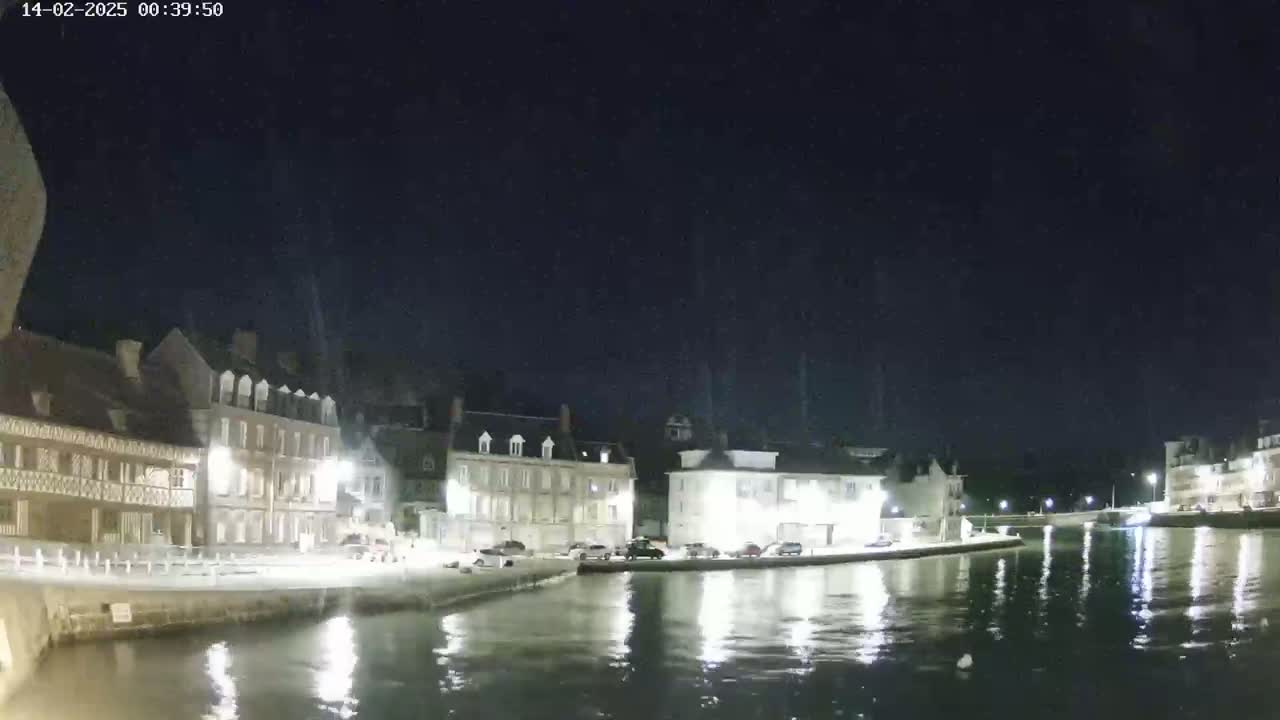 A nighttime view of a calm canal reflecting illuminated buildings along its banks under a clear, dark sky.