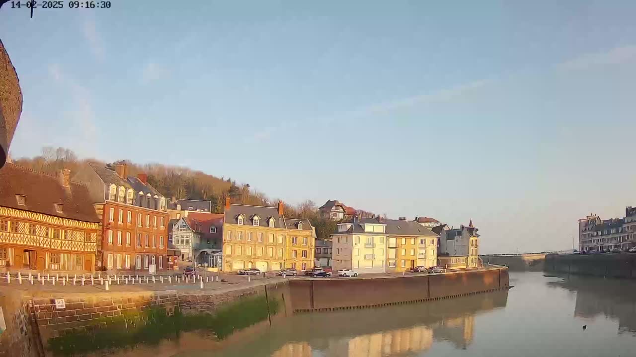 A calm river reflects the sunlit facades of several multi-story buildings, some appearing historical, under a clear, light-blue sky.