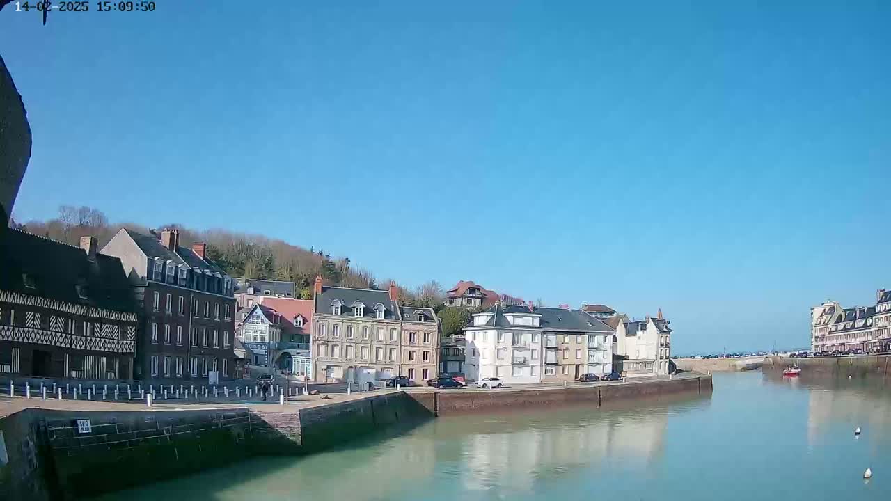 A waterfront town with several multi-story buildings lines a calm waterway under a clear blue sky.