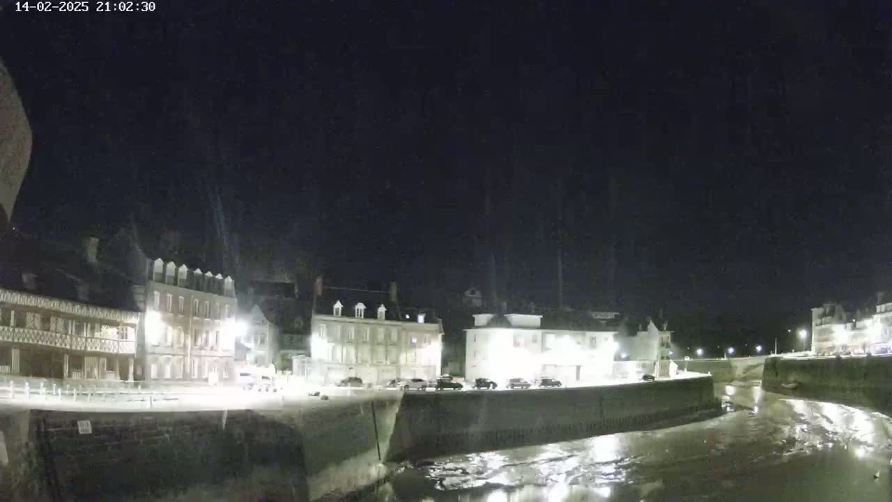 A nighttime view of a waterfront town with several buildings lit up along a waterway at low tide under a clear, dark sky.