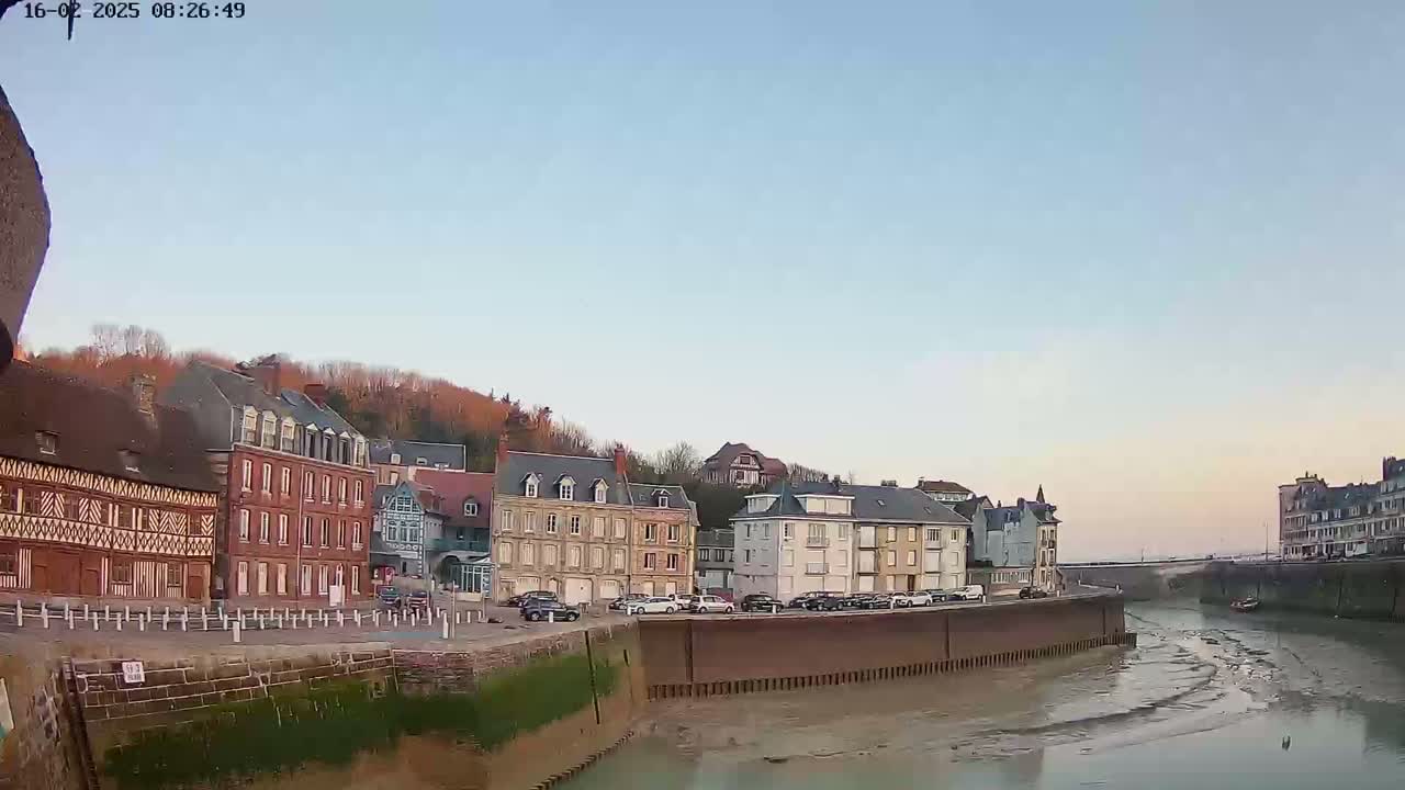 A waterfront town with a mix of architectural styles is shown under a clear, light-blue sky, with a calm waterway showing low tide.