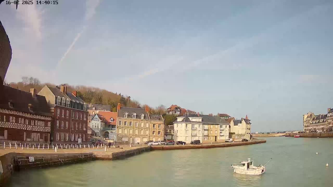 A small white boat floats on calm water in a harbor, lined with multi-story buildings under a mostly clear sky.