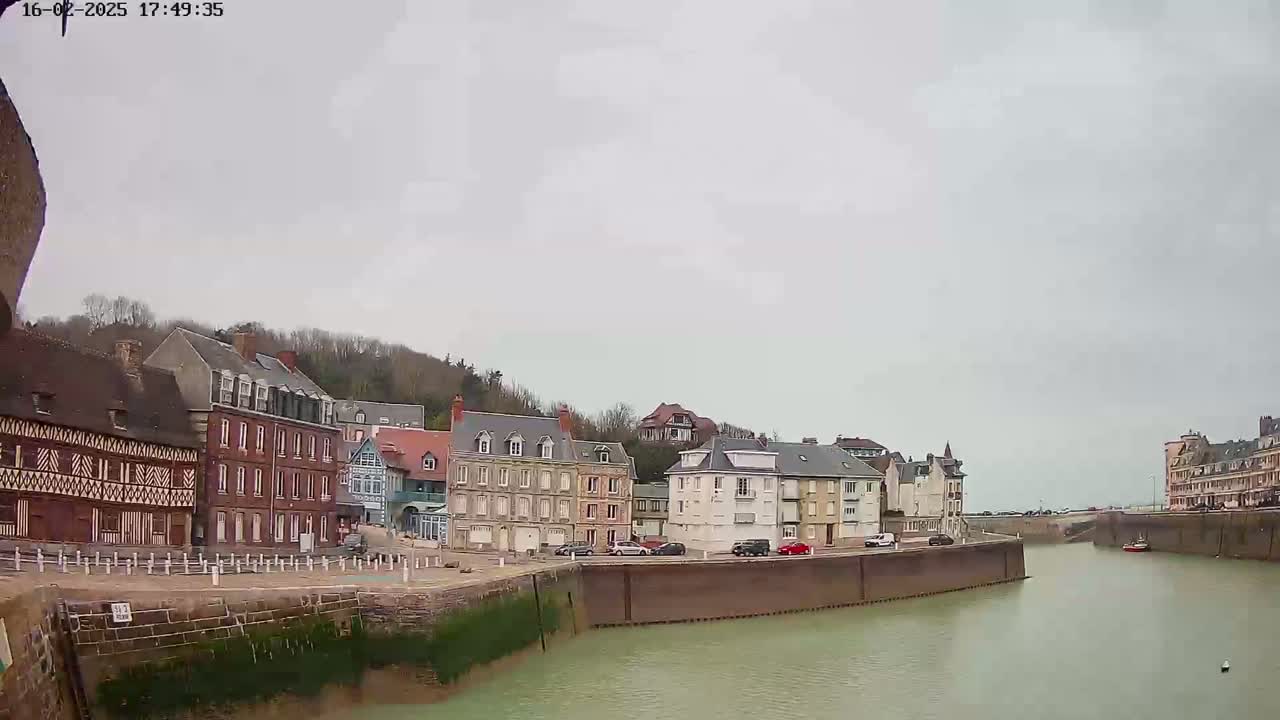 A calm, overcast day reveals a waterfront town with several multi-story buildings lining a canal, some cars parked nearby.